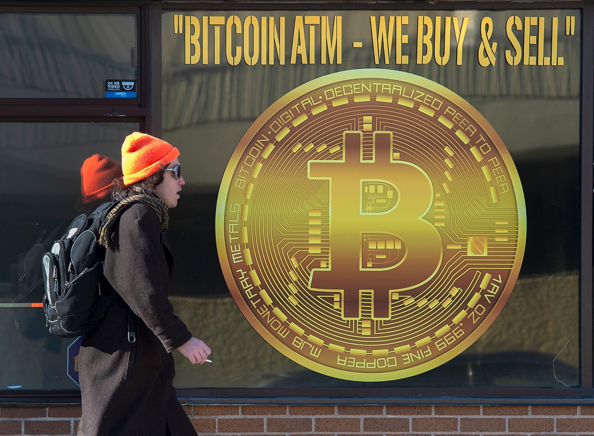 A person wearing an orange hat, long dark coat and backpack walks past a sign advertising bitcoin.