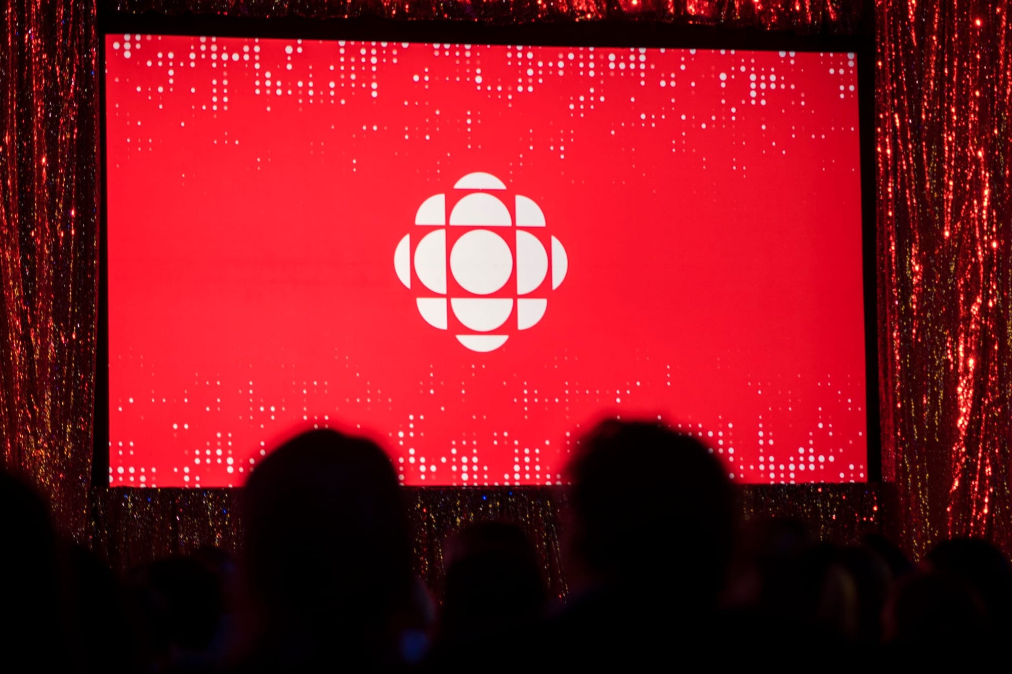 The heads of a small audience are silhouetted in front of a red screen with a projection of the CBC logo.