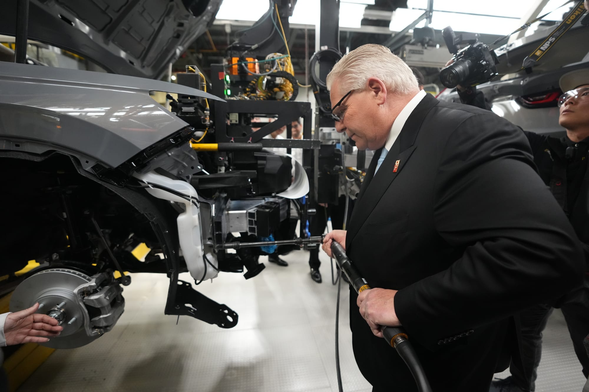 Doug Ford, in a suit and protective glasses, holds a long tool in a car manufacturing facility