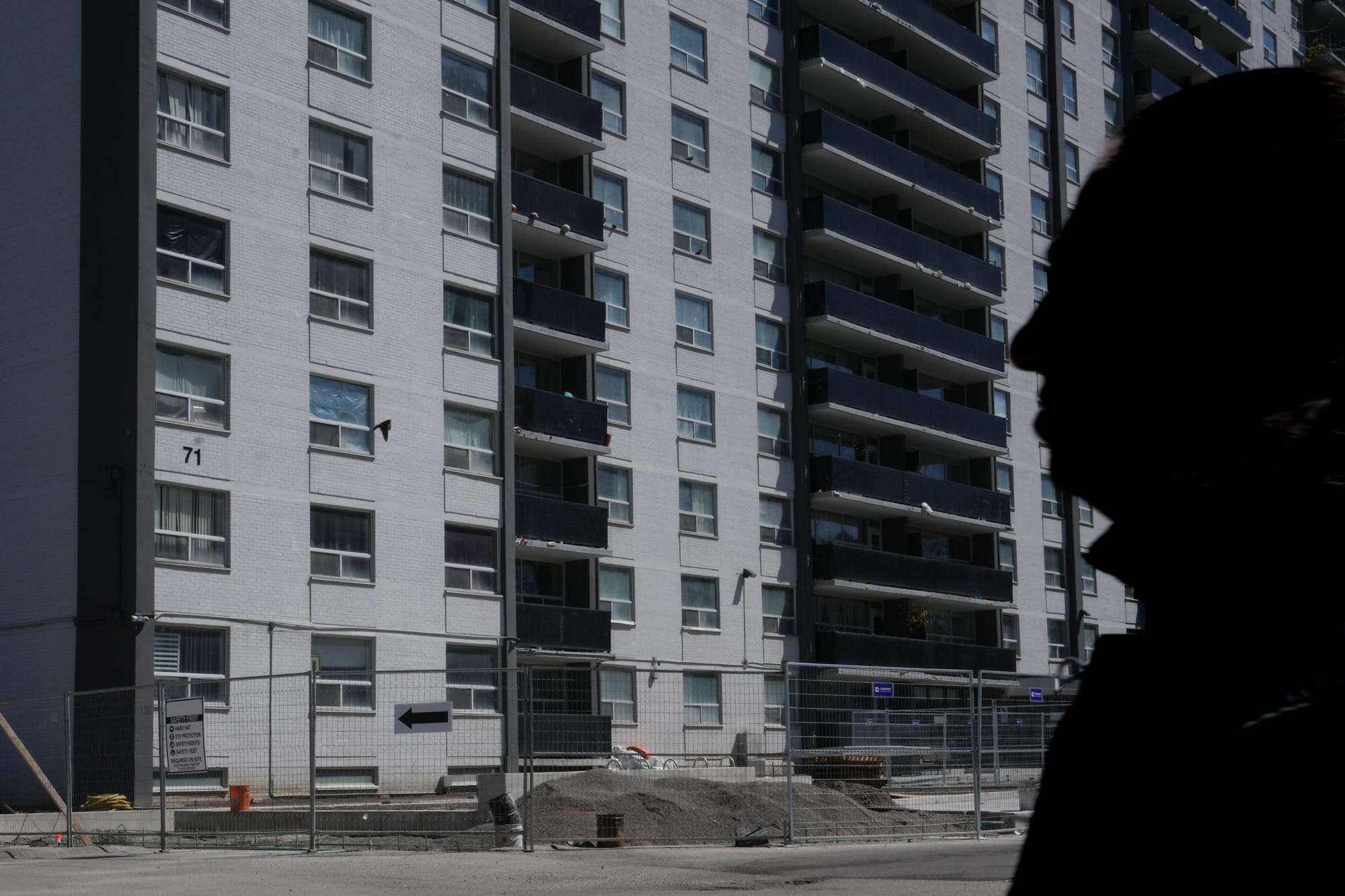 An exterior shot of a grey building. A person's face is in silhouette in the foreground on the right side of the image.