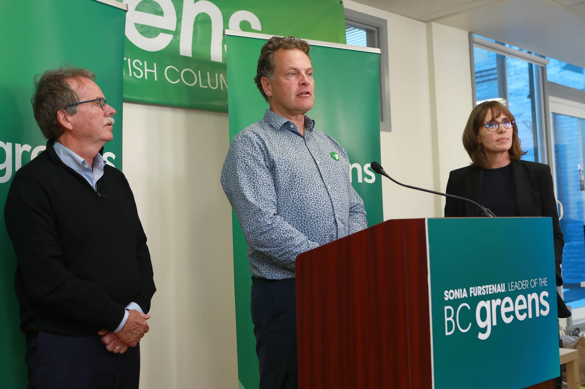 Man in a patterned blue shirt speaks at a podium. He is flanked by another man on the left and a woman in glasses on the right. 