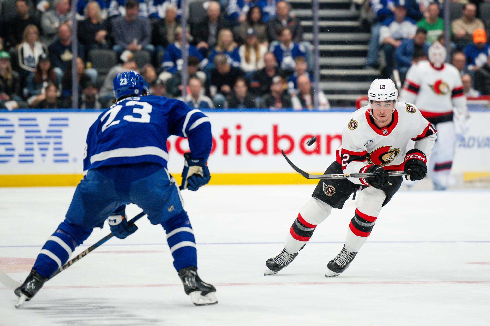 An Ottawa Senators hockey player flips a hockey puck into the air past a Toronto Maple Leafs player.