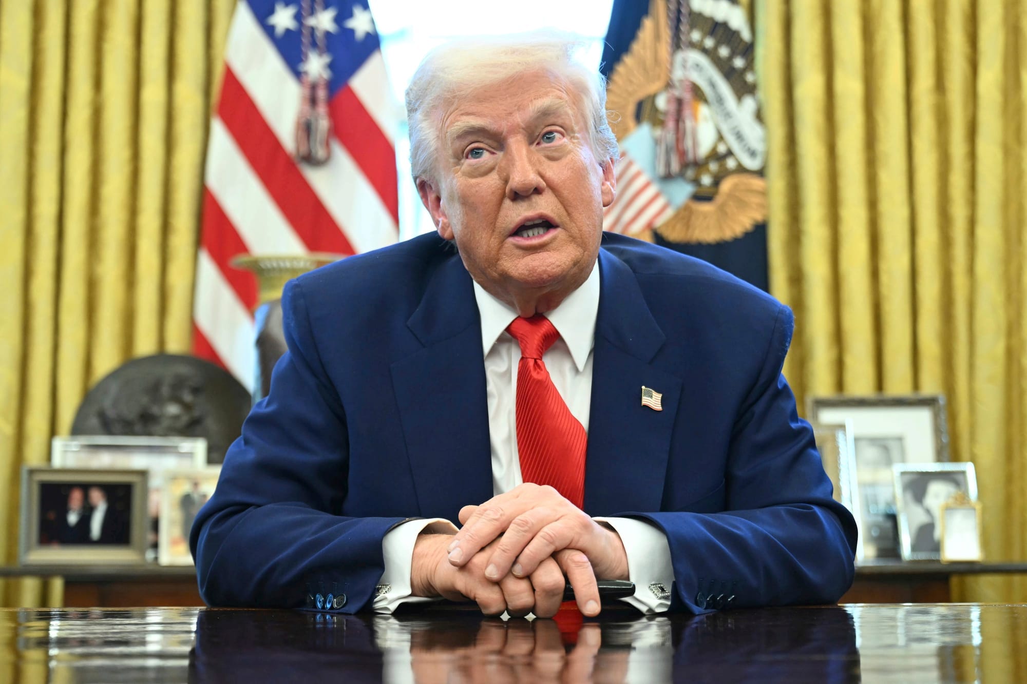Donald Trump is speaking as he wears a blue suit and red tie sitting at his desk in the Oval Office.