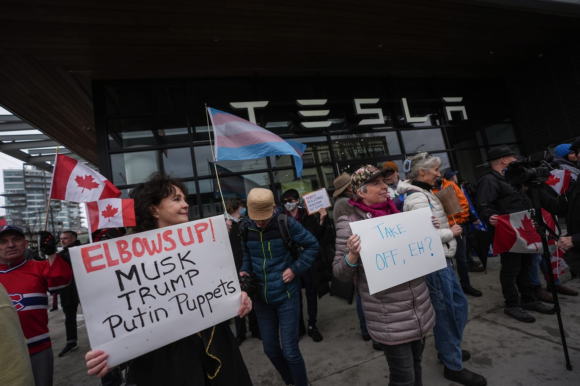 Protestors stand outside a Tesla dealerships while holding transgend pride flags and holding anti-Musk signs