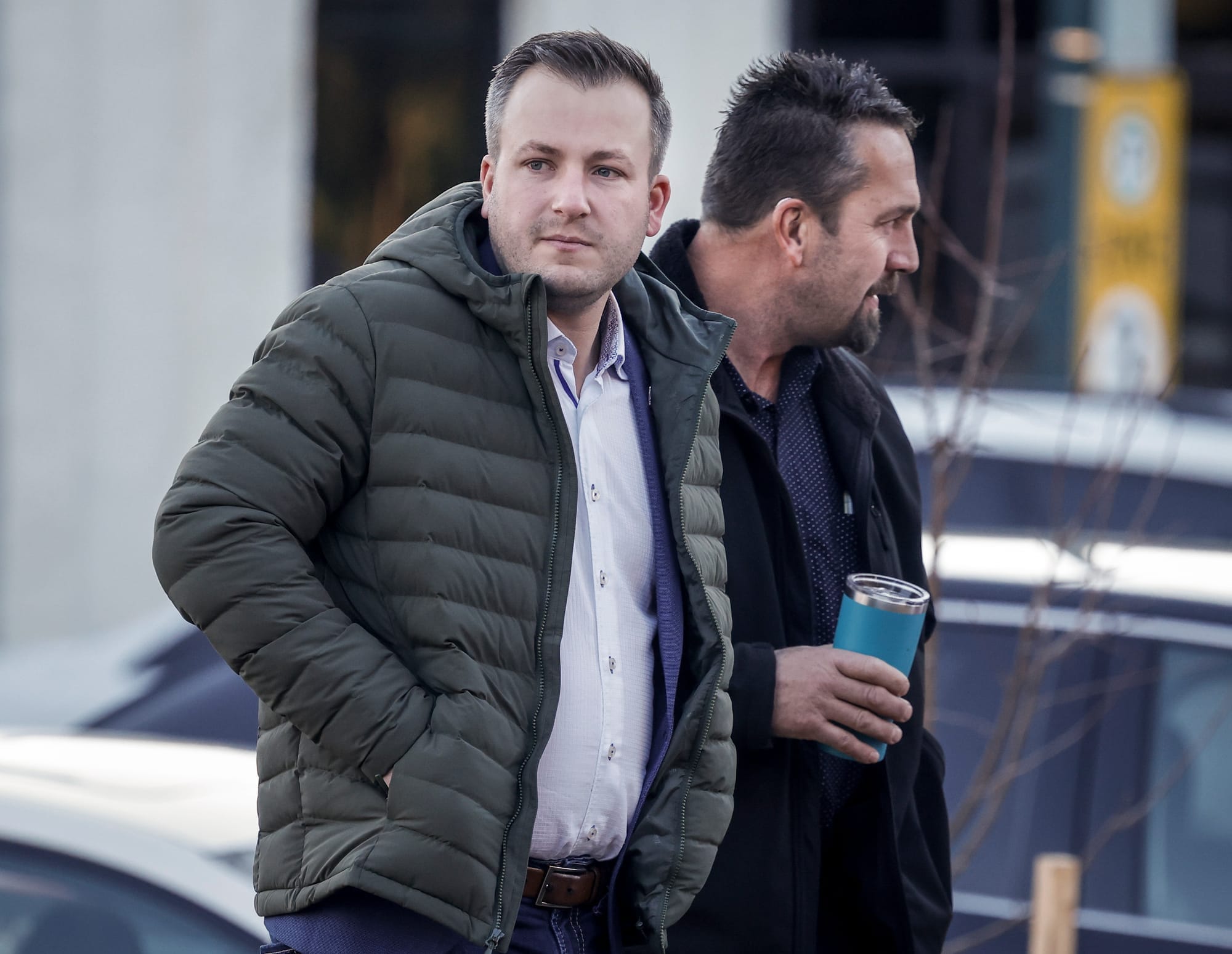 A man in a winter jacket walks next to another man with a goatee holding a portable coffee cup.