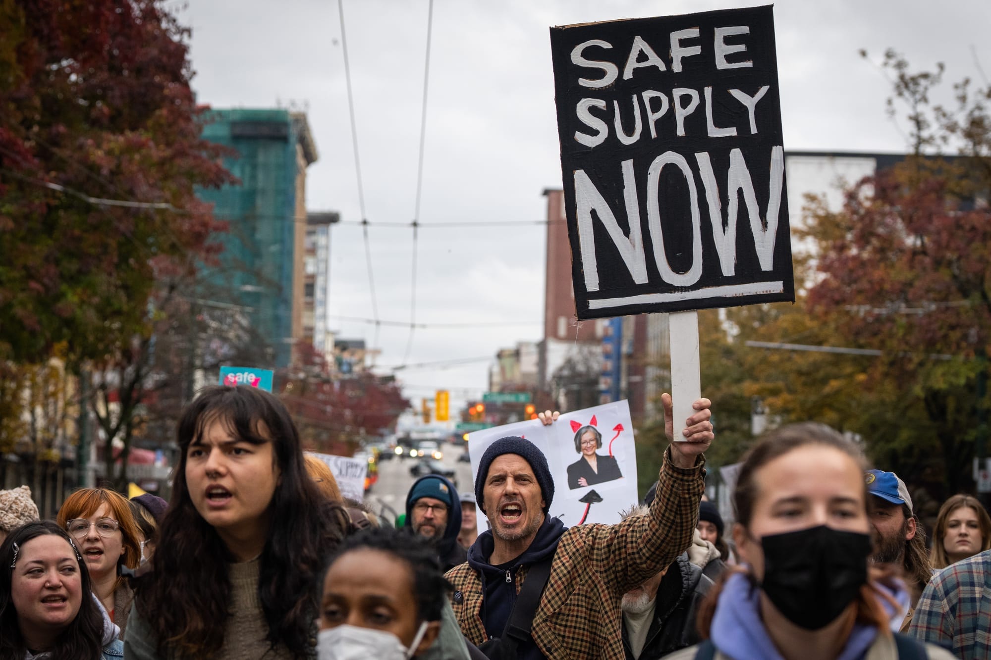 A man holds a sign that says SAFE SUPPLY NOW while in a crowd at a protest in Vancouver. 