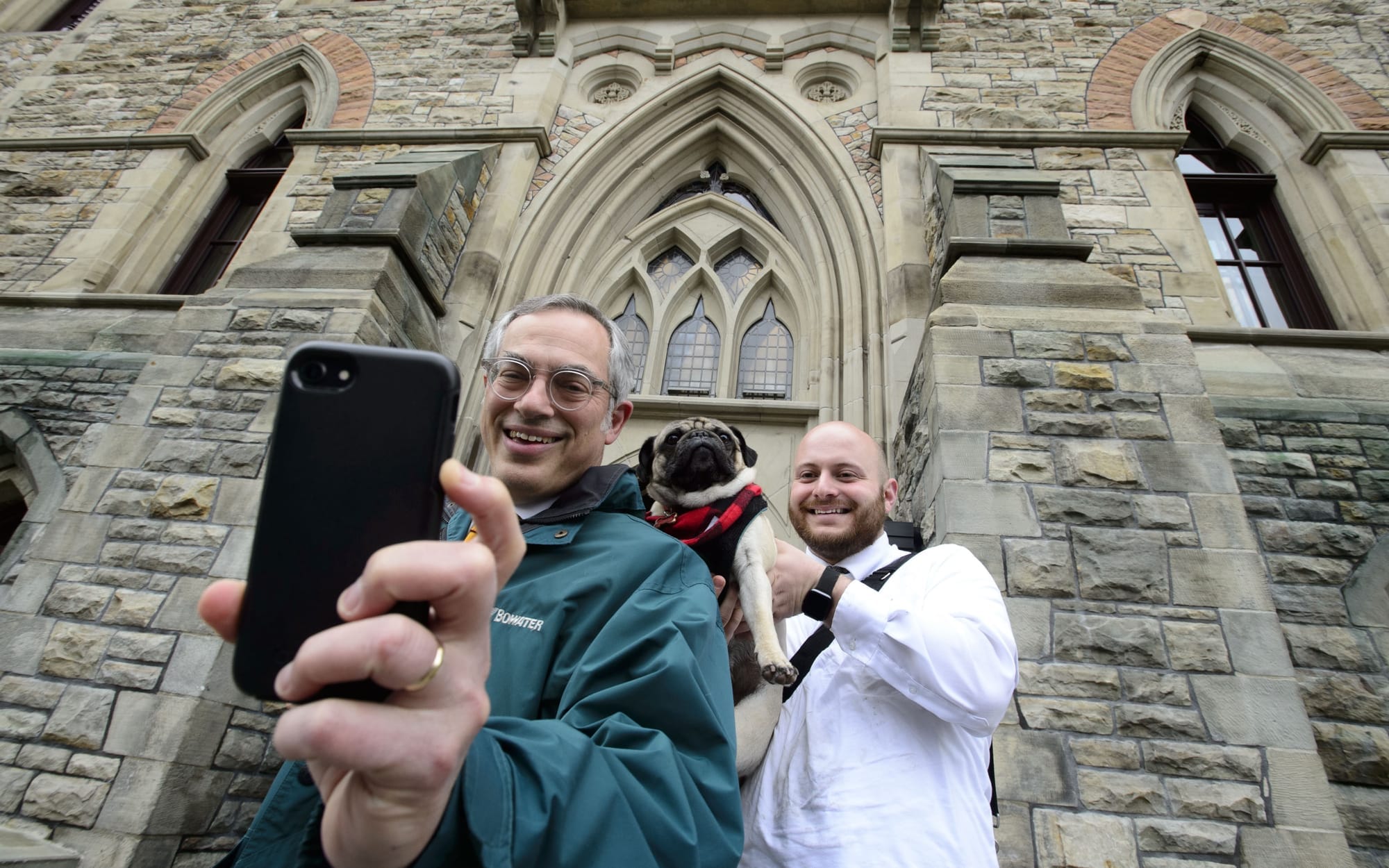 Tony Clement holds a phone for a selfie as a bald man stands behind him holding a pug for the photo.