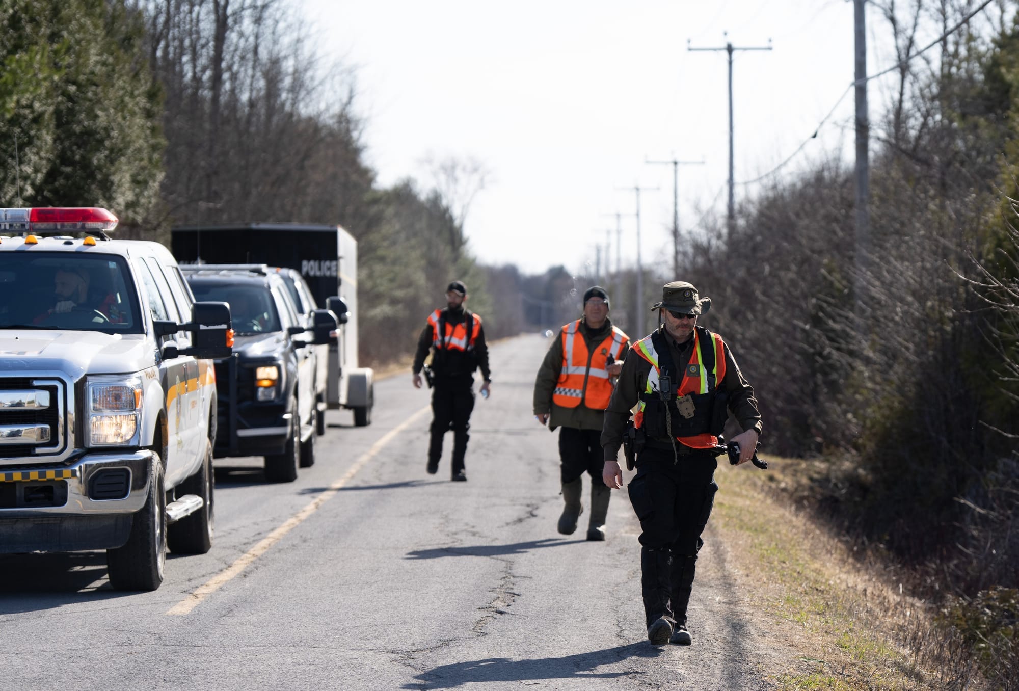 A group of men in orange high visability vests walks along the side of a rural road. 