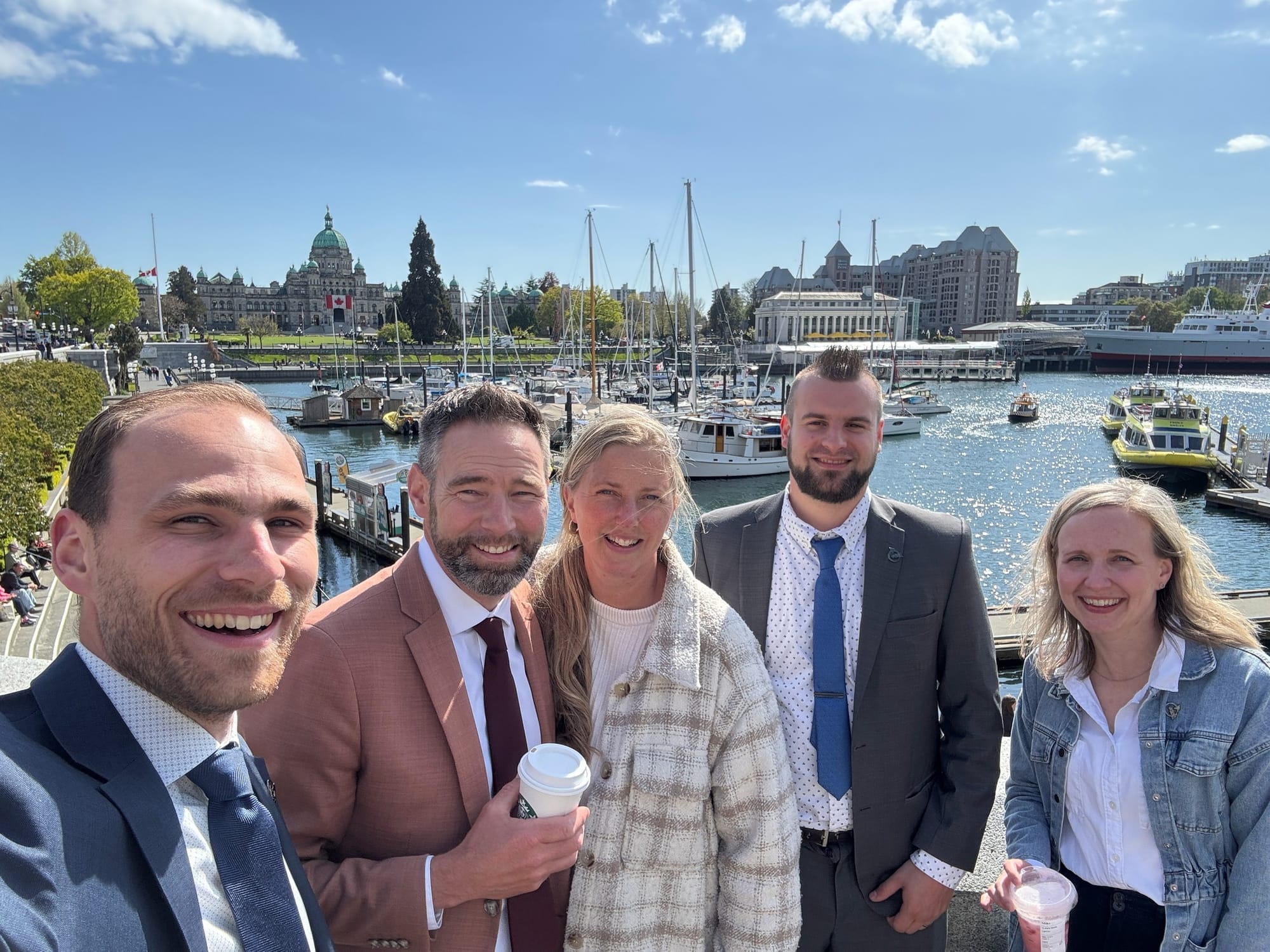 Five people pose for a photo in front of a harbour in Victoria, B.C. with the B.C. Legislature in the background. 