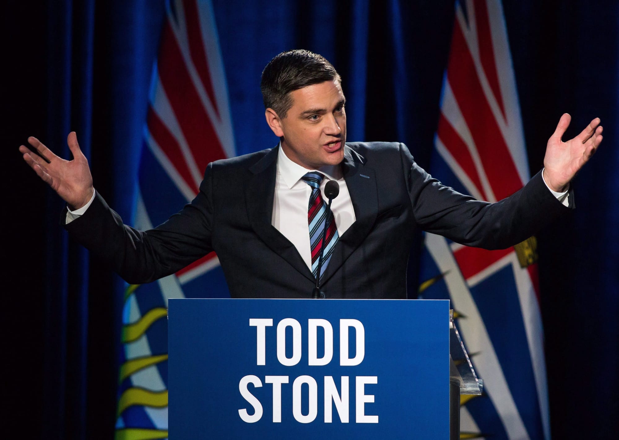 Todd Stone raises his arms in the arm during a speech. He is standing behind a podium that reads 'Todd Stone.