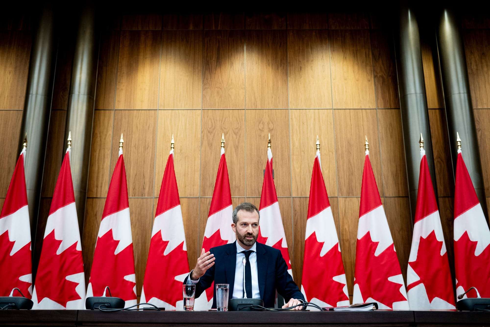 Minister of Government Transformation, Public Works and Procurement Joel Lightbound sits at a long table in front of 10 Canadian flags during a press conference.