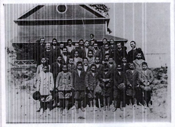 A group of Indigenous children wearing dark suits pose next to two men in clerical collars in a black and white photo.