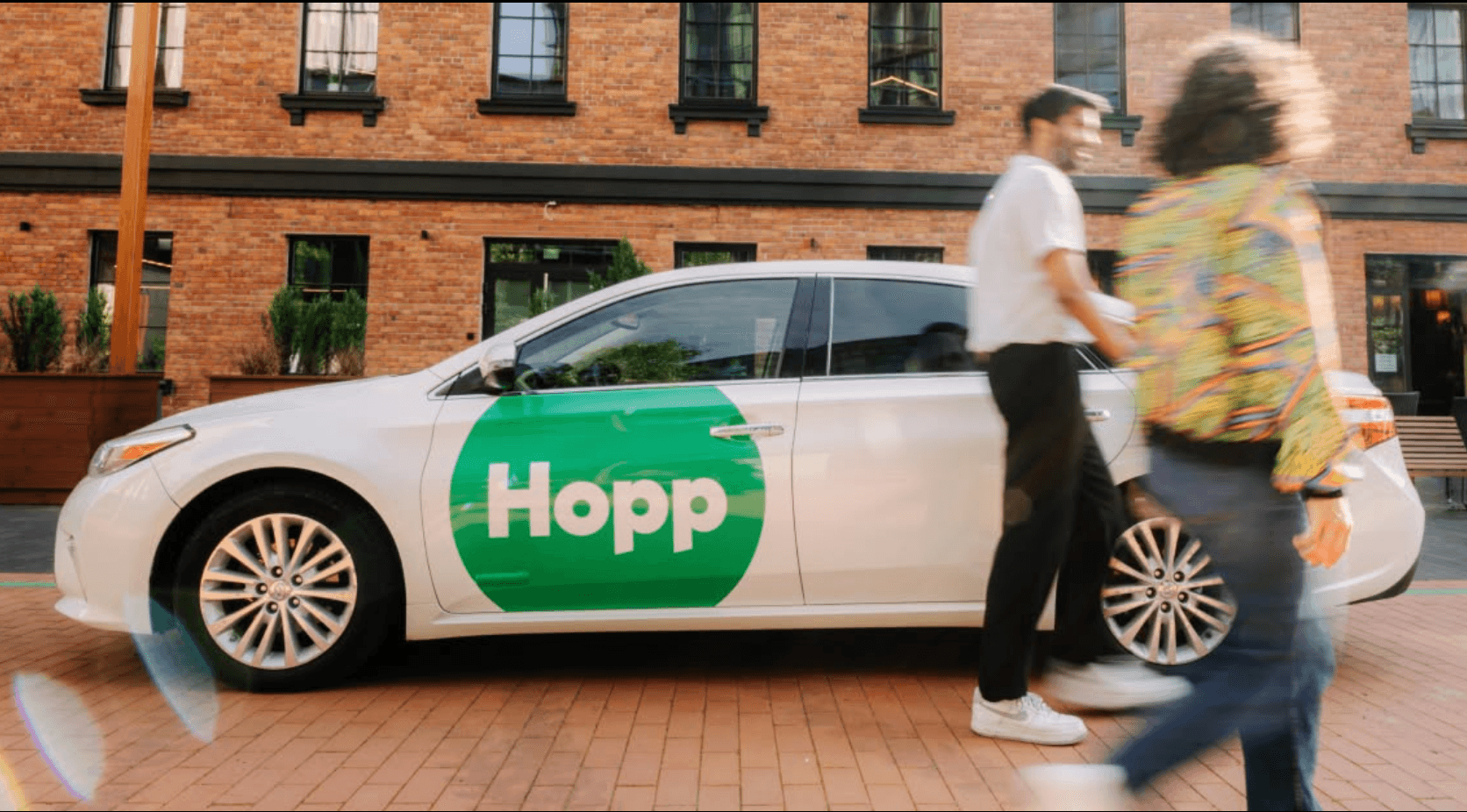 A white sedan with a Hopp logo is parked as people walk by.